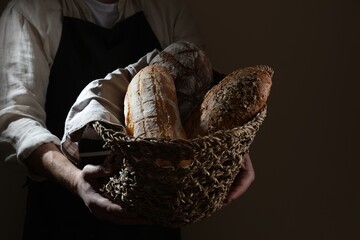 Man holding wicker basket with different types of bread on dark brown background, closeup