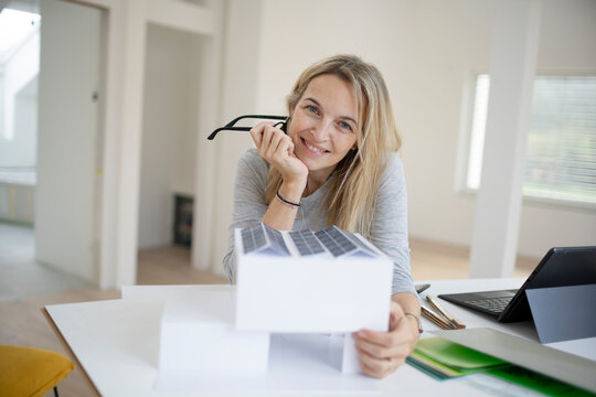 pretty blonde architect with black eyeglasses is standing at a desk and looking at a model of a house with photovoltaic system, solar system, in a loft, office