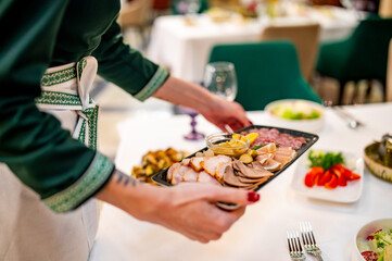 A waiter elegantly serves a platter of assorted deli meats and pickles at a well-set dining table. The scene exudes sophistication, with the waiter in a green shirt and white apron