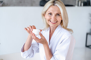 50 years old woman in white bathrobe holding jar with moisturizer cream