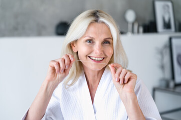 Smiling mature woman in white bathrobe clean teeth using dental floss