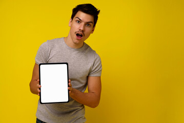 Young caucasian shocked man 25th holds big pad computer with mock up white screen isolated on yellow background studio portrait. modern technology concept