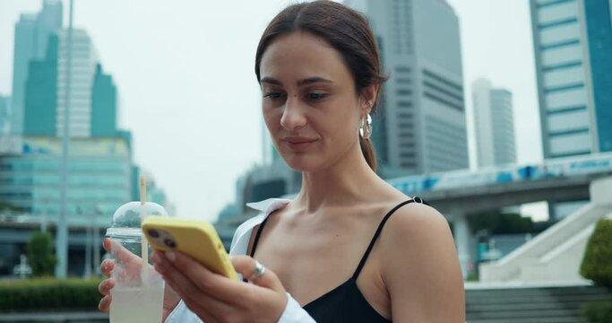 Woman stands against backdrop of Bangkok's modern skyscrapers drinking water using phone close-up. Bangkok symbol of new technology and futuristic architecture Bangkok bustling Thai capital.