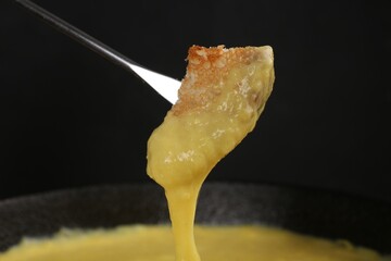 Dipping piece of bread into fondue pot with tasty melted cheese against dark gray background,...