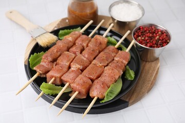 Wooden skewers with cut raw marinated meat, spices and basting brush on white tiled table
