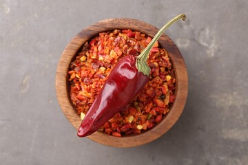 Chili pepper flakes in bowl and pod on grey table, top view