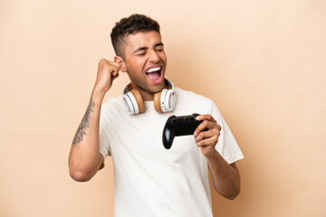 Young Brazilian man playing with a video game controller isolated on beige background celebrating a victory © luismolinero