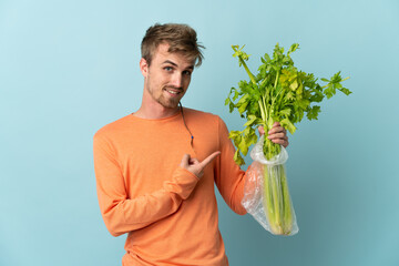 Young blonde man holding a celery isolated on blue background and pointing it