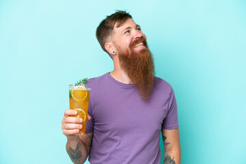 Redhead man with long beard holding a cocktail isolated on blue background looking up while smiling