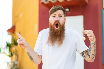 Redhead man with beard holding home keys at outdoors surprised and pointing side