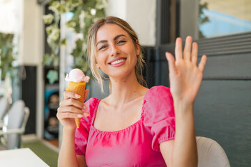 Young blonde woman with a cornet ice cream at outdoors saluting with hand with happy expression