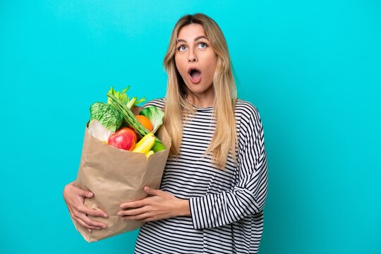 Young Uruguayan woman holding a grocery shopping bag isolated on blue background looking up and with surprised expression