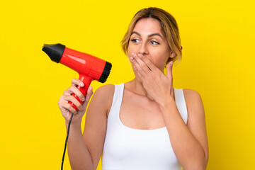 Young Uruguayan woman holding a hairdryer isolate don yellow background with surprise and shocked...