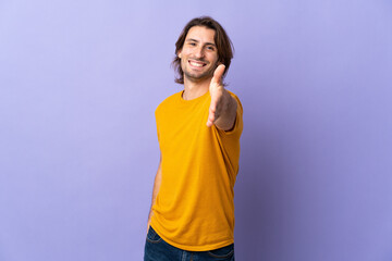 Young handsome man isolated on purple background shaking hands for closing a good deal