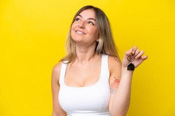 Young Rumanian woman holding home keys isolated on yellow background looking up while smiling
