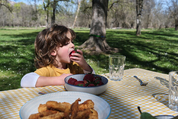 A young boy is eating a strawberry while sitting at a table with a bowl of fruit