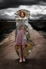 woman is standing on a road wearing a silver and gold rescue blanket