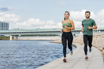 Couple jogging by the river, cityscape in background, sunny day, both wearing fitness attire.