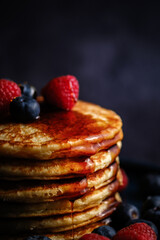 Stacked pancakes with raspberries and blueberries and maple syrup against a dark background.