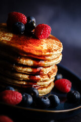 Stacked pancakes with raspberries and blueberries and maple syrup against a dark background.