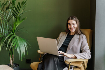 Happy professional woman working on a laptop in a cozy corner with a green plant beside her.