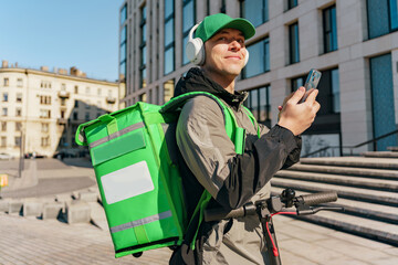 Smiling delivery man with green backpack checking smartphone in urban setting, headphones on.