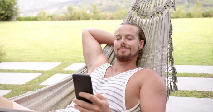 A young Caucasian male relaxes in a hammock in the backyard at home, smartphone in hand