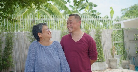An senior retirement Asian married couple sharing a joyful moment standing in a sunny garden, smiling at the camera. Scene is happy and relaxed