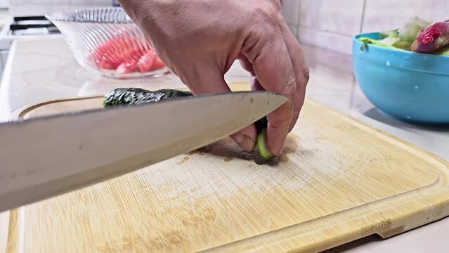 Caucasian hands cutting short green cucumber on bamboo cutting board at domestic kitchen, wide angle close-up with slow motion. Real life user-generated content.