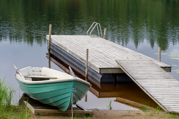 Wooden dock and a green rowing boat anchored at shore. Lake at background with trees reflecting...