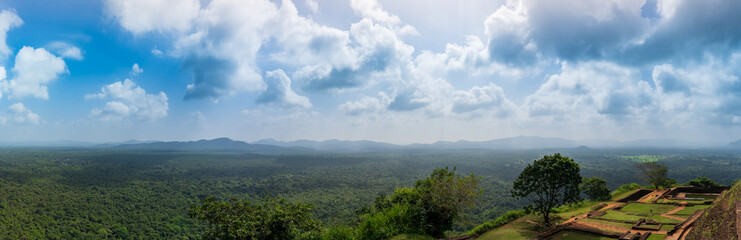 An afternoon panoramic view from the top of Sigiriya Rock, overlooking a vast green forest with distant mountain ranges under a blue sky.