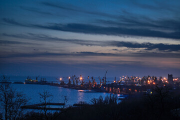 Twilight descends on busy port with cranes silhouetted against dusk sky. Maritime industrial hub transitions to night, highlighting harbor activity and urban backdrop with emerging city lights.