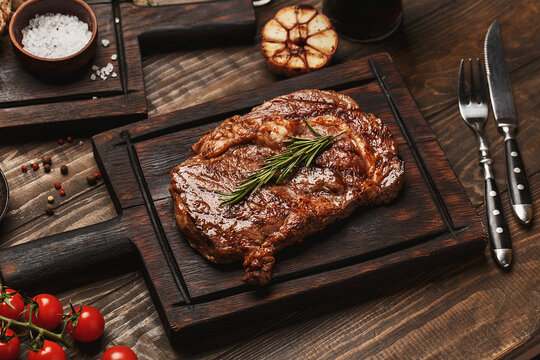 Grilled ribeye beef steak served on wooden board with rosemary, grilled garlic, fork and knife on wooden table