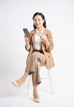 Full body image of young Asian businesswoman sitting and using phone on white background