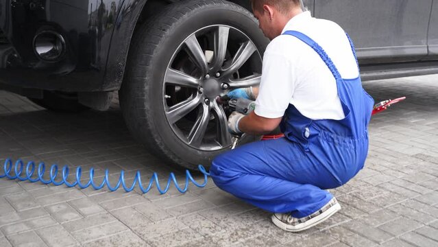 Auto mechanic in blue uniform tightens lug nuts on car wheel using pneumatic air gun in workshop. Vehicle maintenance tech fastens tire with impact wrench, performs pit stop service.