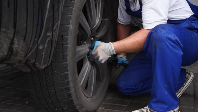 Auto mechanic in blue uniform installs tire using torque wrench at service station. Professional tightens lug nuts on car wheel for secure fit. Vehicle maintenance, accuracy in automotive industry.