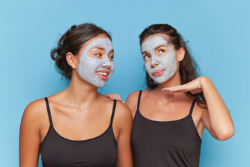 Two girlfriends pose over blue background shoulder to shoulder, clay cosmetic mask on their faces, skincare concept, copy space