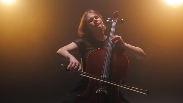 Concentrated female musician holding cello and creating wonderful music while performing on theatre scene. Attractive woman playing musical instrument while closing eyes and enjoying concert.