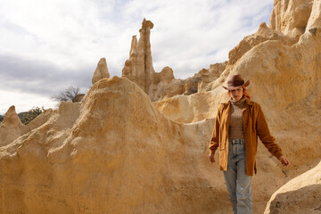 A woman wearing a brown jacket and a hat walks through a desert landscape. The scene is desolate...