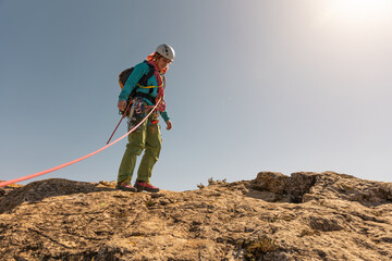 A woman is standing on a rocky hill, wearing a backpack and a red harness. She is looking down,...