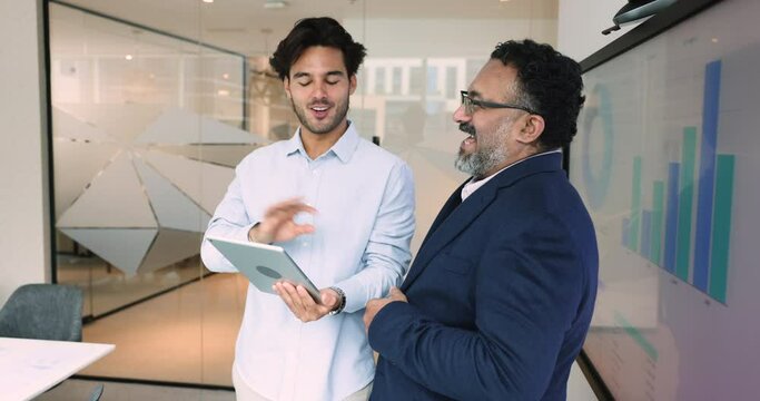 Laughing Indian and Mexican businessmen collaborate using digital tablet device. CEO and employee discuss financial strategy, review charts, share ideas, consider task together in friendly environment