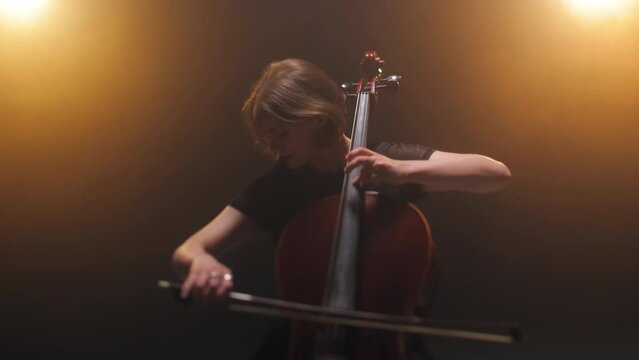 Attractive woman performing while holding elegant cello and expressing lyrical feeling with facial expression. Famous female musician playing musical instrument on scene lit with yellow lighting.