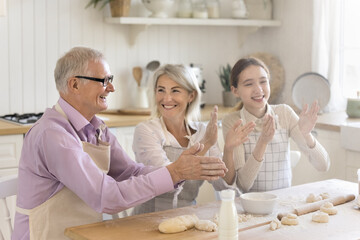 Multi-generational family laughing, having fun, joking, cooking together, clap hands, enjoy cooking process, make homemade dough for holiday pastries spend weekend time together in modern cozy kitchen