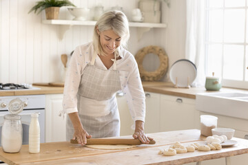 Beautiful middle-aged housewife cooking in modern cozy kitchen. Smiling mature woman in apron flattening dough with rolling-pin, baking sweet buns alone at home. Culinary, homemade food preparation