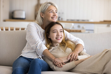Fototapeta na wymiar Cheerful adolescent girl spends leisure time with mature mother at home, sit resting on sofa together, enjoy tender moments, show love and care. Happy family of two generations hugging and daydreaming