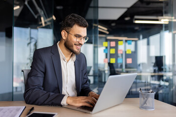 A professional young businessman is smiling while working on his laptop in a well-lit modern office...