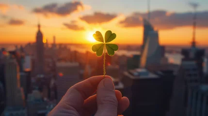 Deurstickers A foreground illustration of a hand holding a four-leaf clover as a symbol of good luck and the symbolism of the traditional St. Patrick's Day holiday in the background of a city at sunset © Zoran