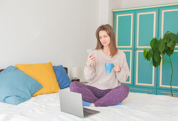 Happy woman sitting on the bed and working with computer and smartphone at home with cup of tea - 760093237