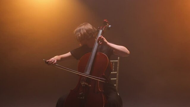 Woman with short hair sitting on stage and looking up while playing emotional song. Concentrated female musician playing cello and enjoying classic melody during concert in theater hall.