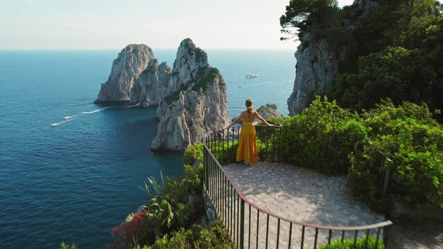 Woman in yellow overlooking the stunning seascape of Capri Island from a cliffside. Iconic Faraglioni sea stacks surrounded by the beauty of nature.